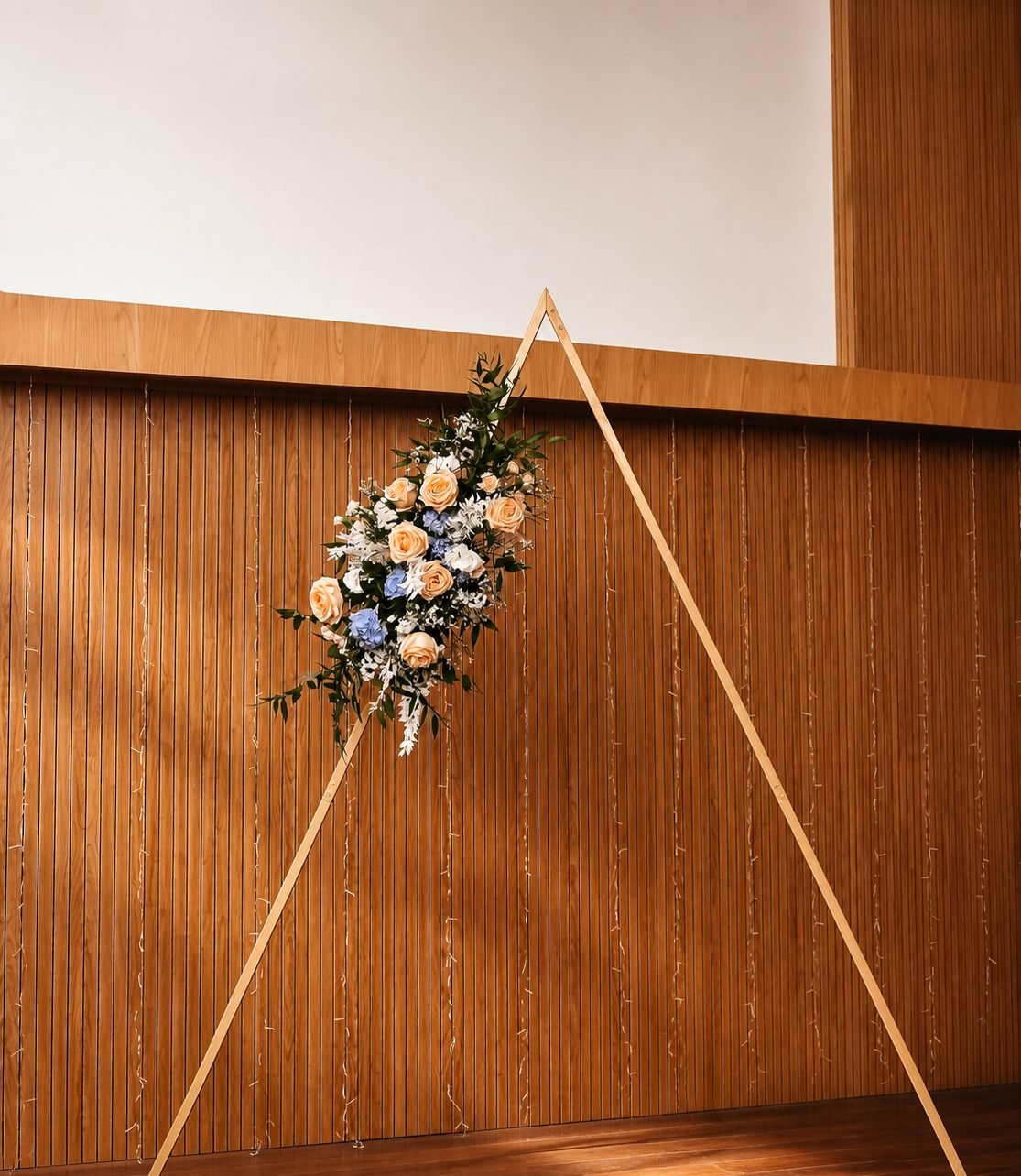 Triangular wooden ceremony frame with peach, white and blue flowers against a warm wood-slatted wall with fairy lights