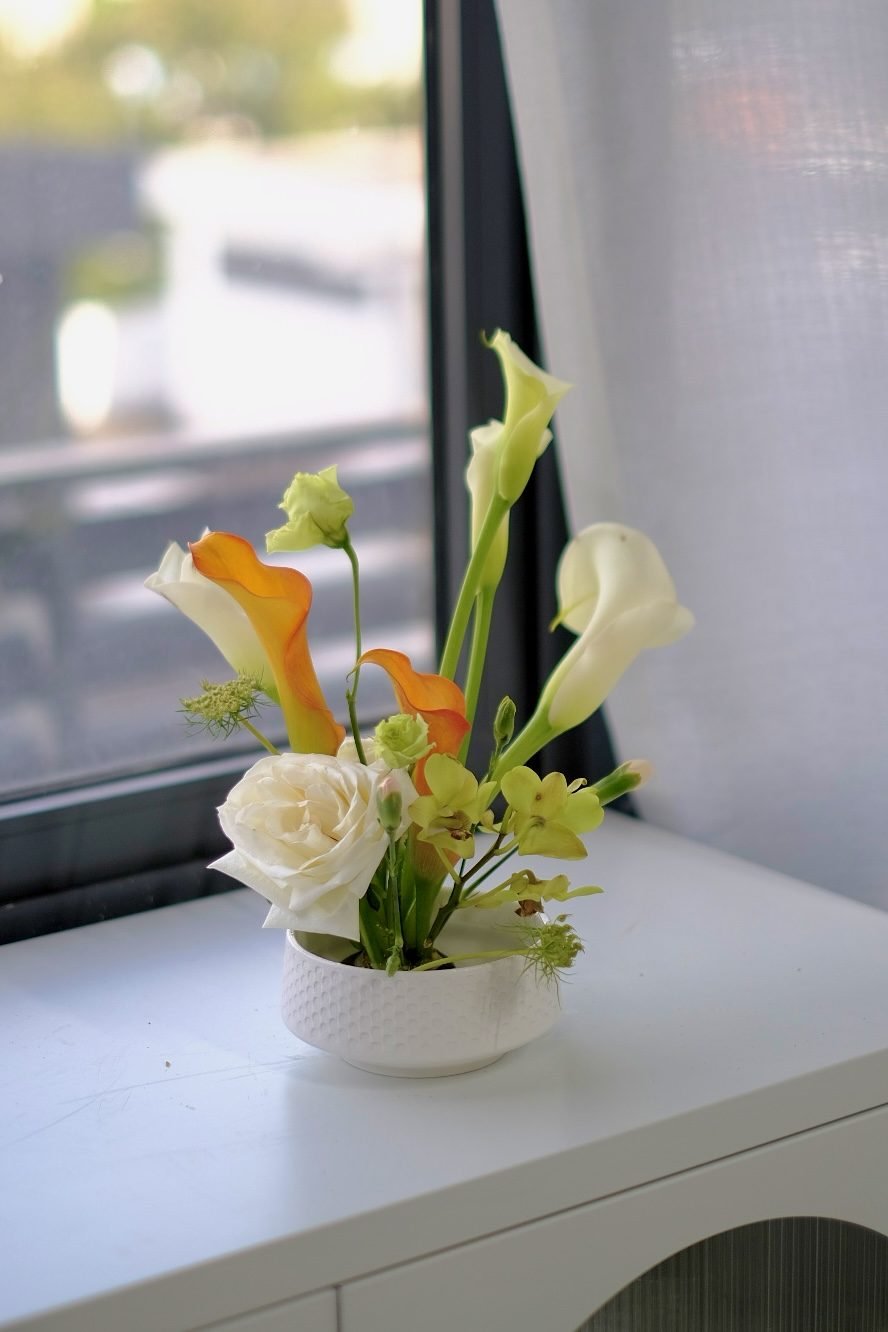 Small individual arrangement of white calla lilies, an orange calla and a white rose by a window