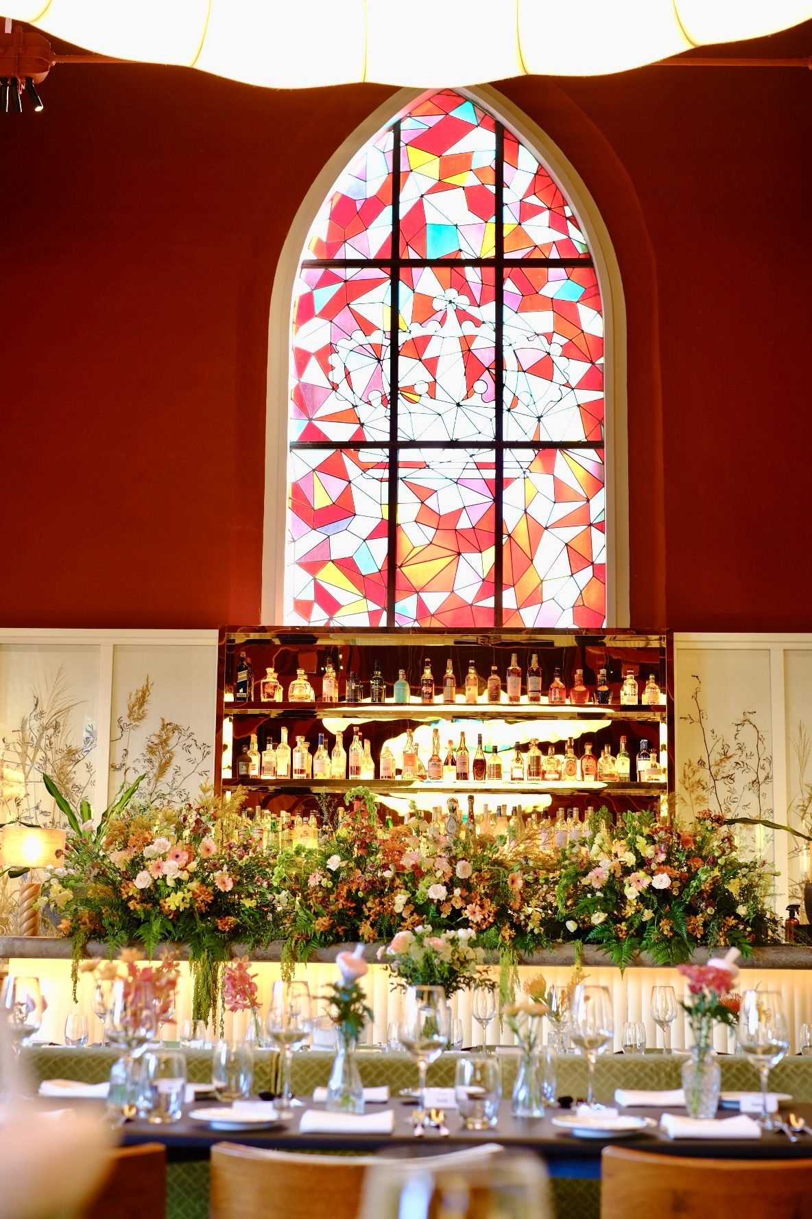 Abundant green and blush flowers lining a restaurant bar beneath an arched stained-glass window