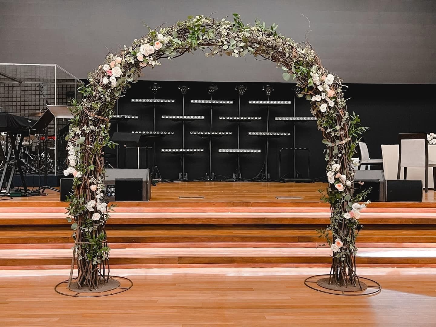Woven twig arch draped with greenery and blush and white flowers in front of a dark stage