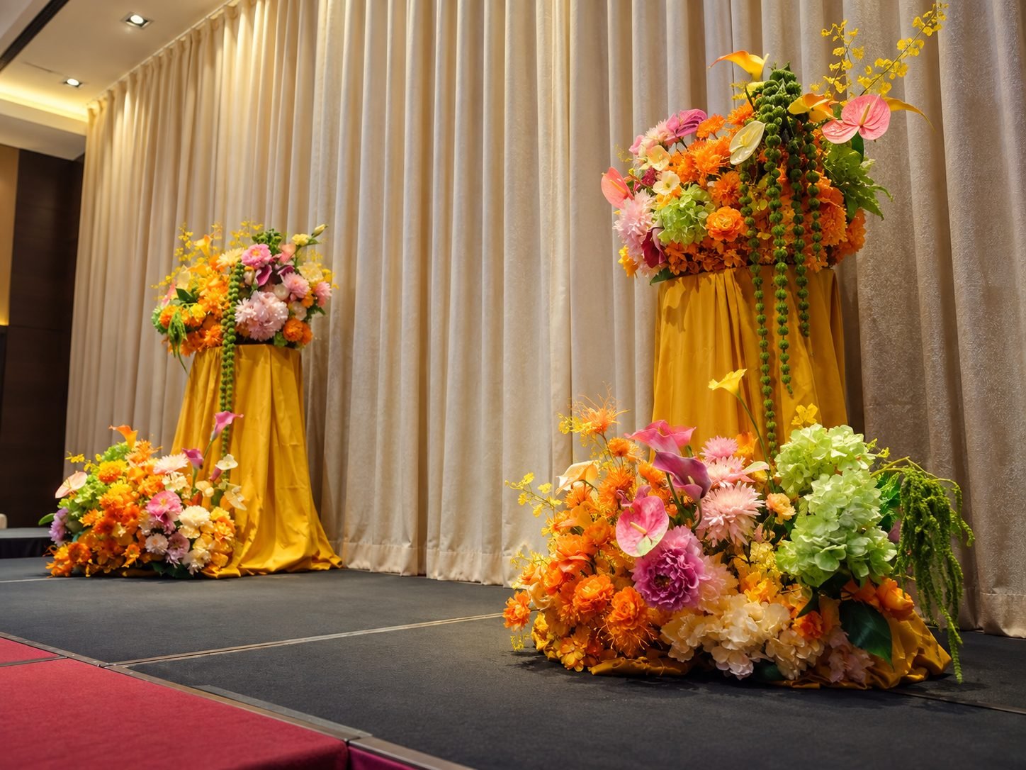 Two tall mustard-draped pedestals topped with orange, pink and green arrangements on a ballroom stage