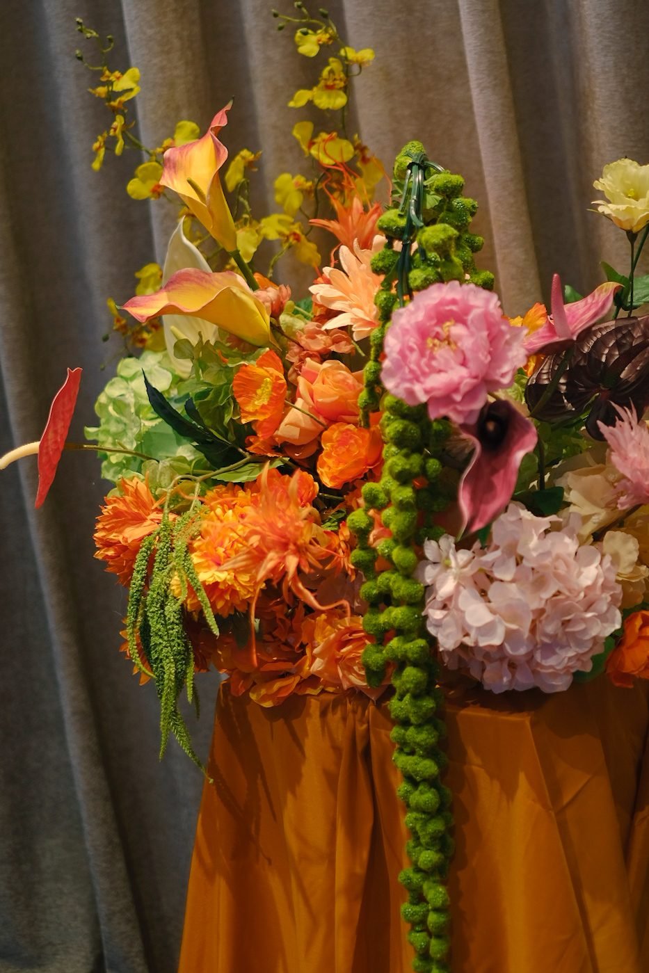 Close detail of a pedestal arrangement with a pink peony, orange dahlias, calla lilies and hanging green beads