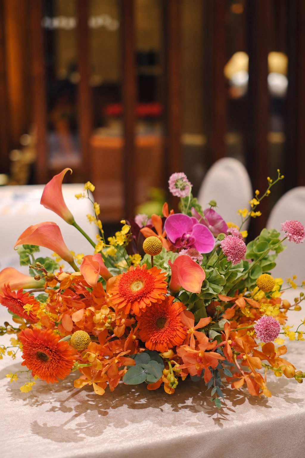 Close-up of a reception table arrangement with orange gerberas, a fuchsia orchid and yellow calla lilies