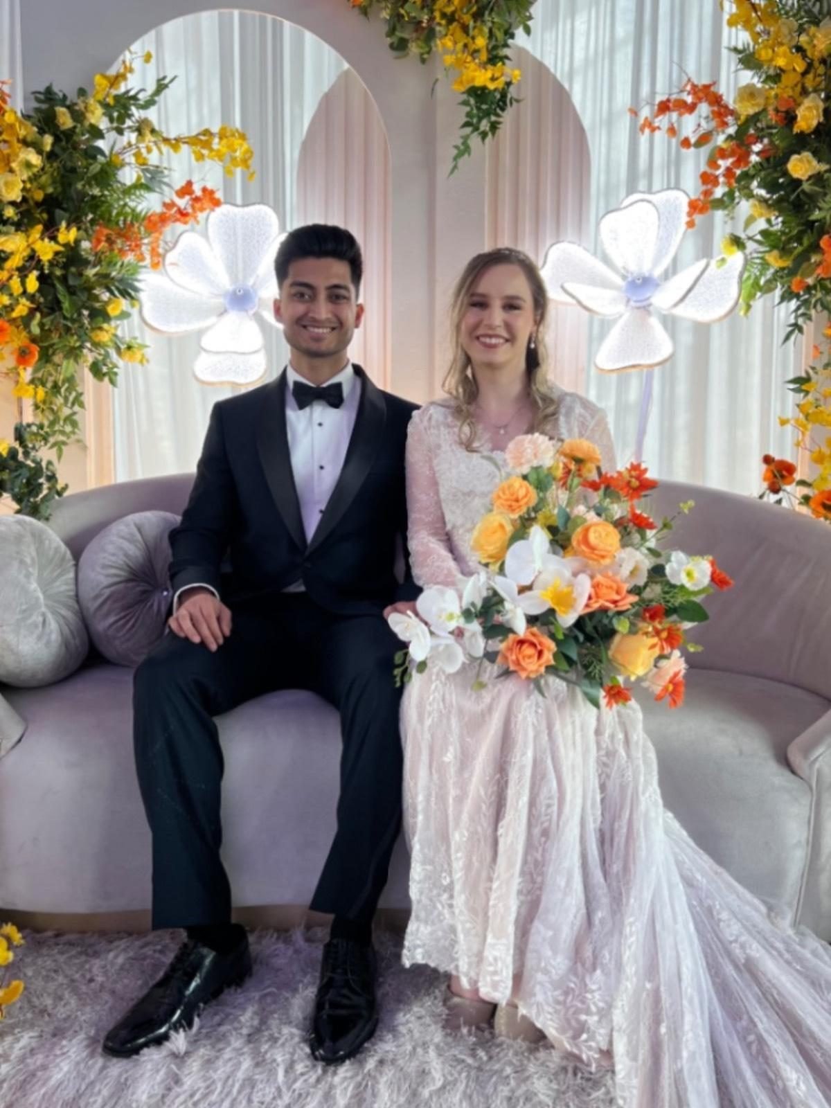 Bride and groom seated on a blush velvet sofa against an arched floral backdrop