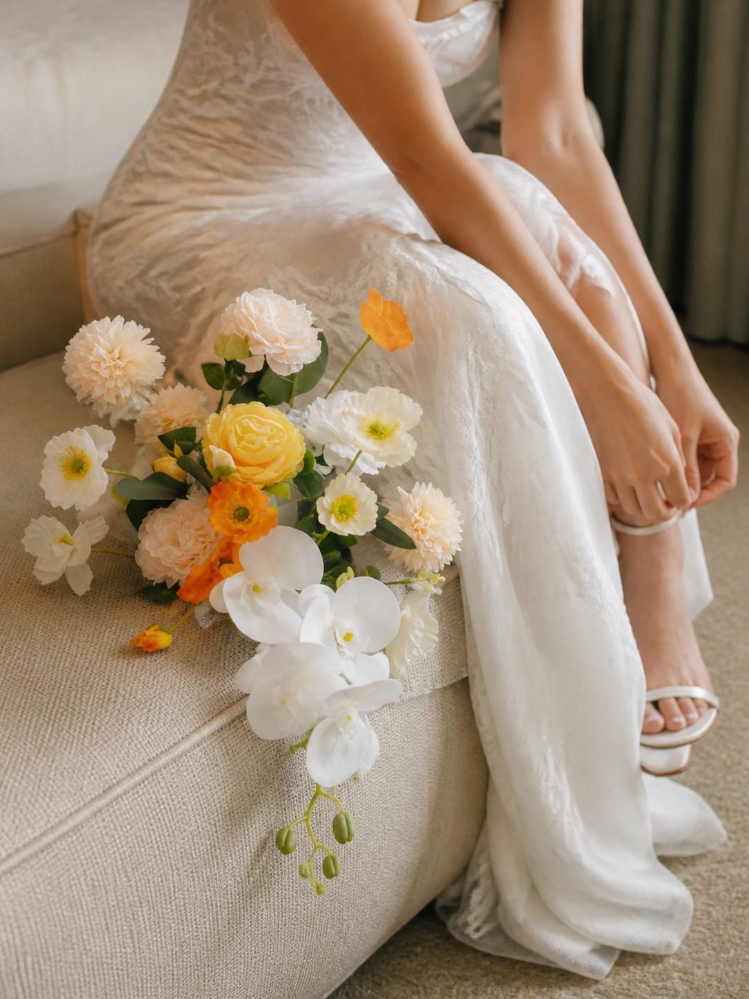 Bride in a lace dress resting a peach, yellow and white bouquet on a sofa beside her