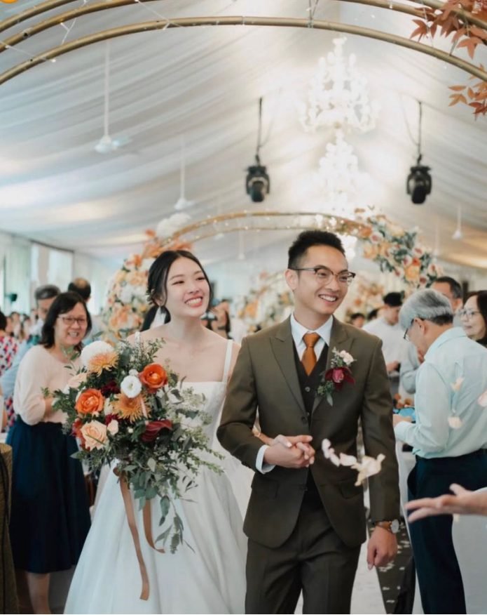 Bride and groom walking arm-in-arm down the aisle beneath a gold arch in a draped marquee