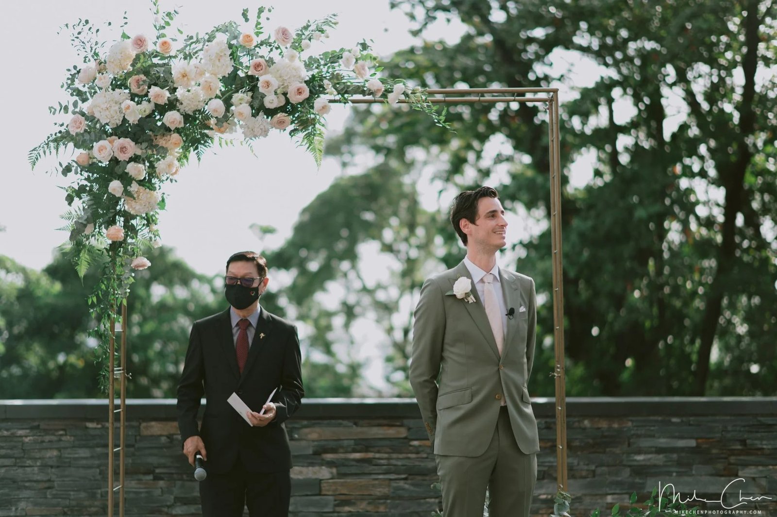 Groom in a sage suit standing under a brass floral arch with blush and white flowers, green trees behind