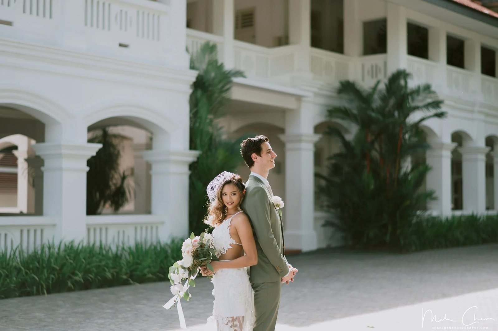 Bride and groom standing back to back in front of a white colonial building with arched verandahs and palms