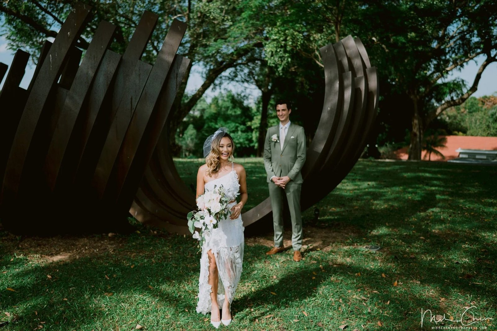 Bride and groom on a lawn in front of a large curved dark metal garden sculpture