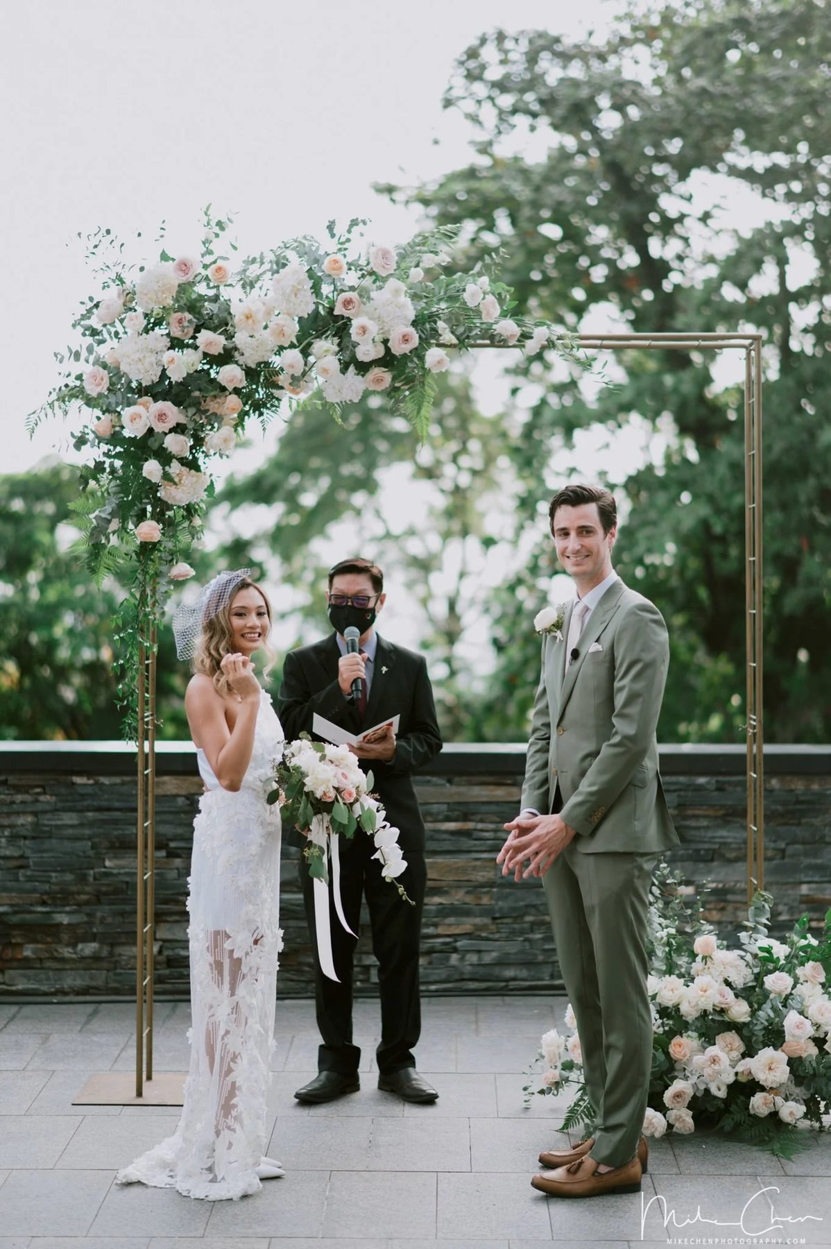 Bride in a white lace gown carrying a white and blush trailing bouquet, groom in a sage green suit beneath a garden arch
