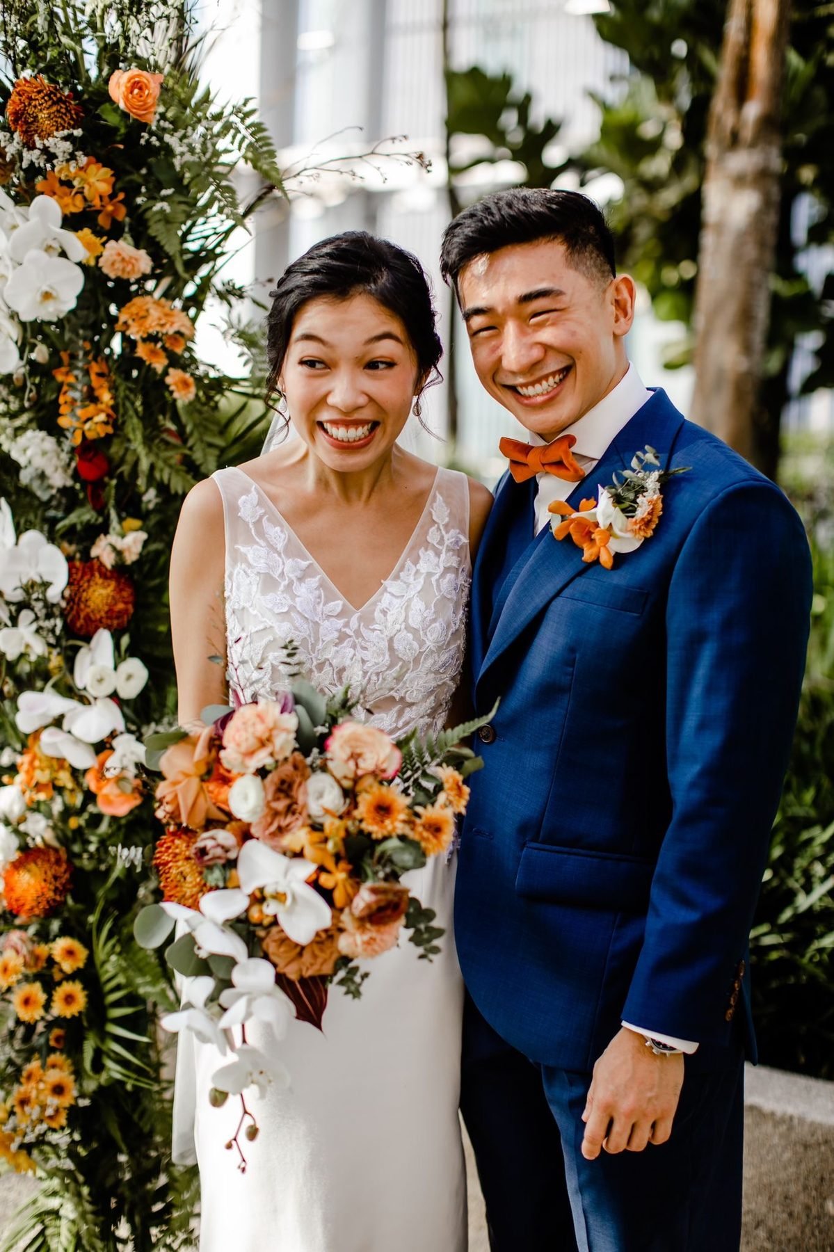Bride and groom laughing, the bride holding a warm-toned cascading bouquet beside a floral pillar