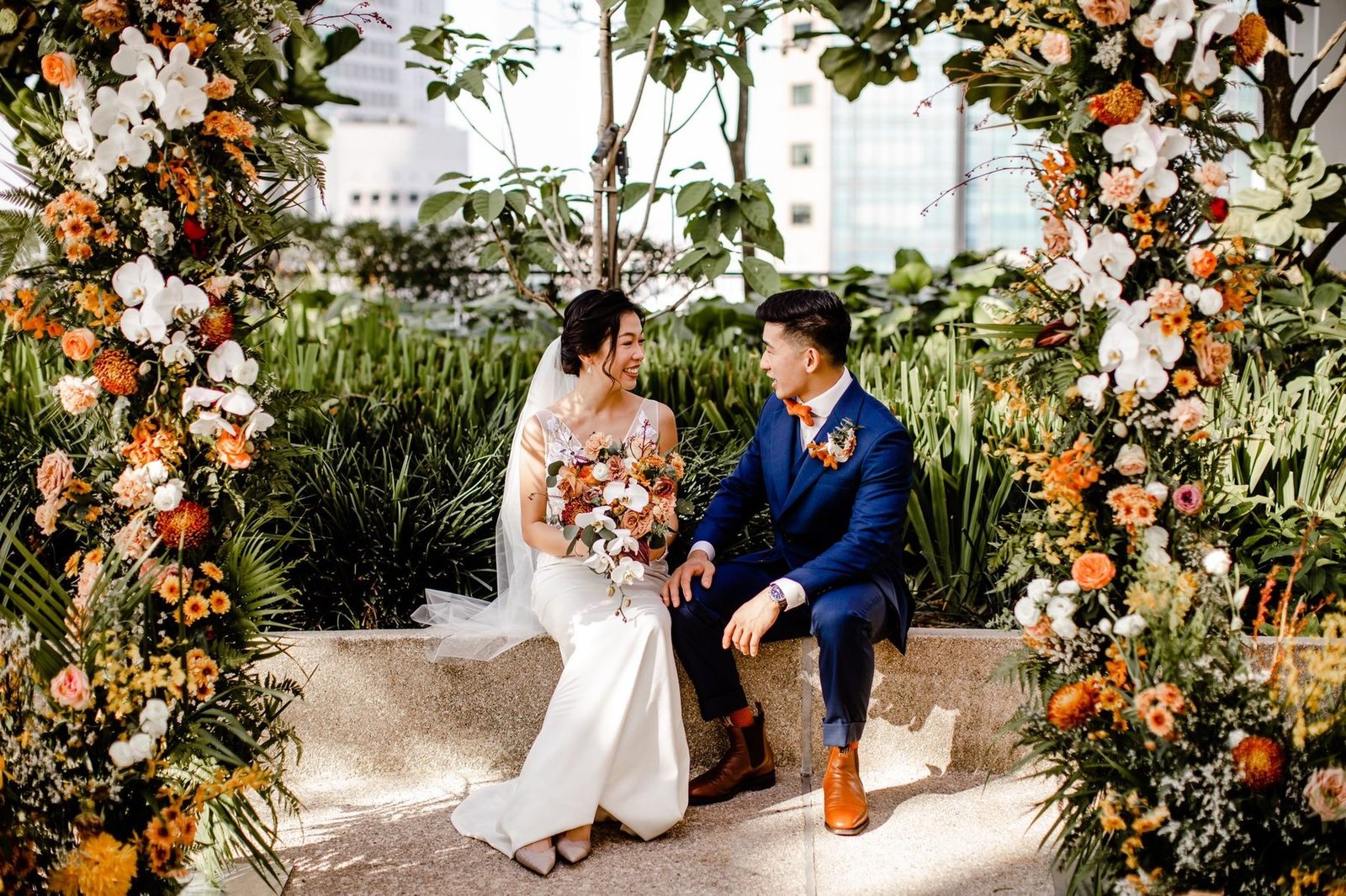 Bride and groom seated on a low ledge framed by two orange and white floral pillars with greenery and a city skyline behind