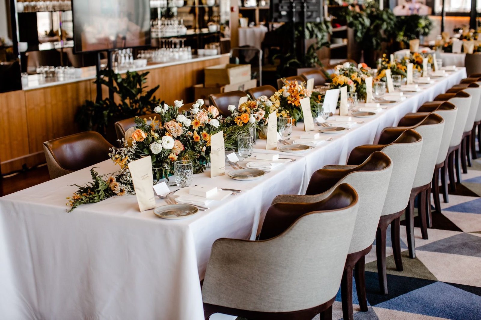 Long white reception banquet table lined with low orange and white floral arrangements in a restaurant