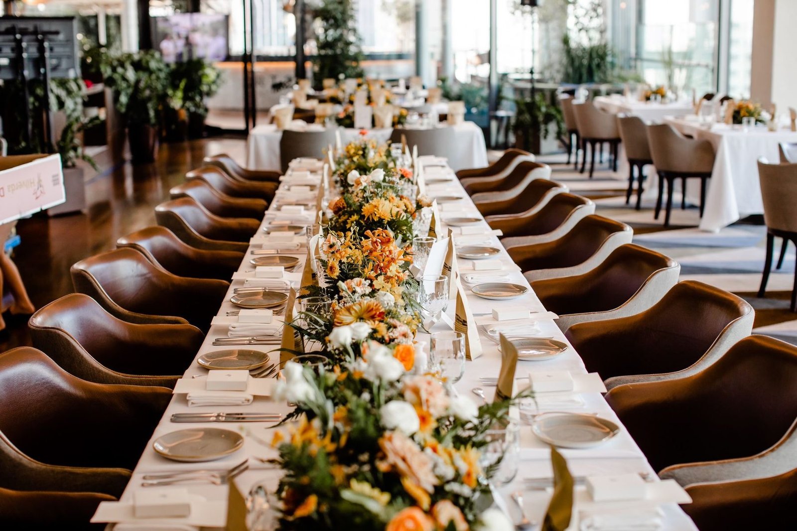 View down a long banquet table with a continuous orange, yellow and white floral runner and leather chairs on either side