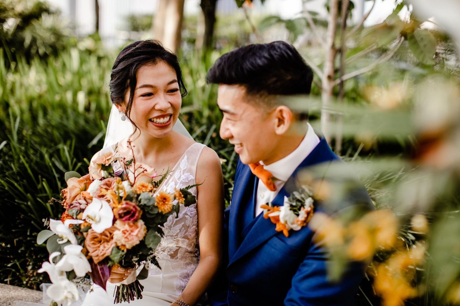 Bride holding a warm-toned bouquet laughing beside the groom in a navy suit, garden greenery behind