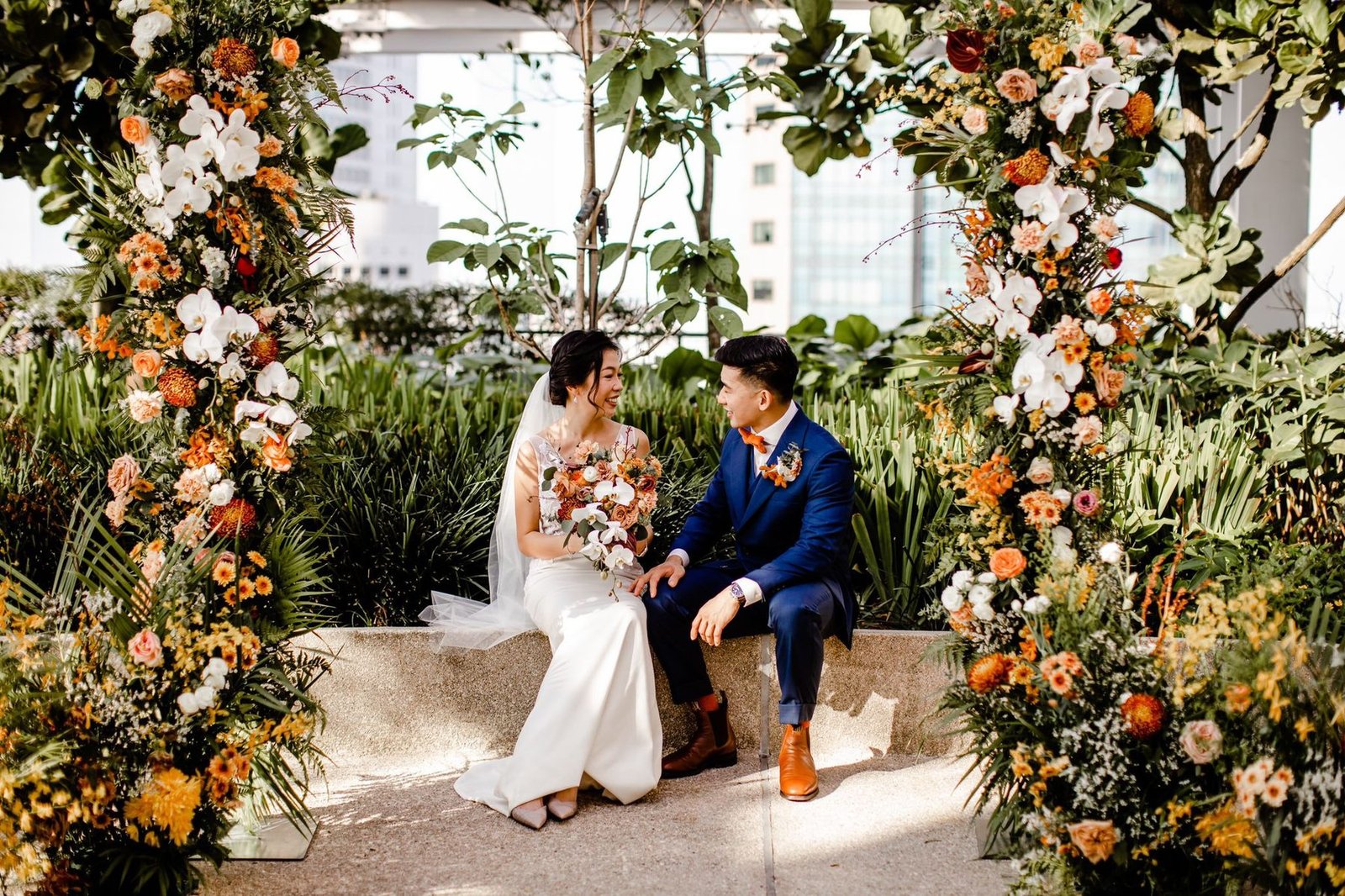 Bride and groom seated on a stone ledge between two orange and white floral pillars on a rooftop