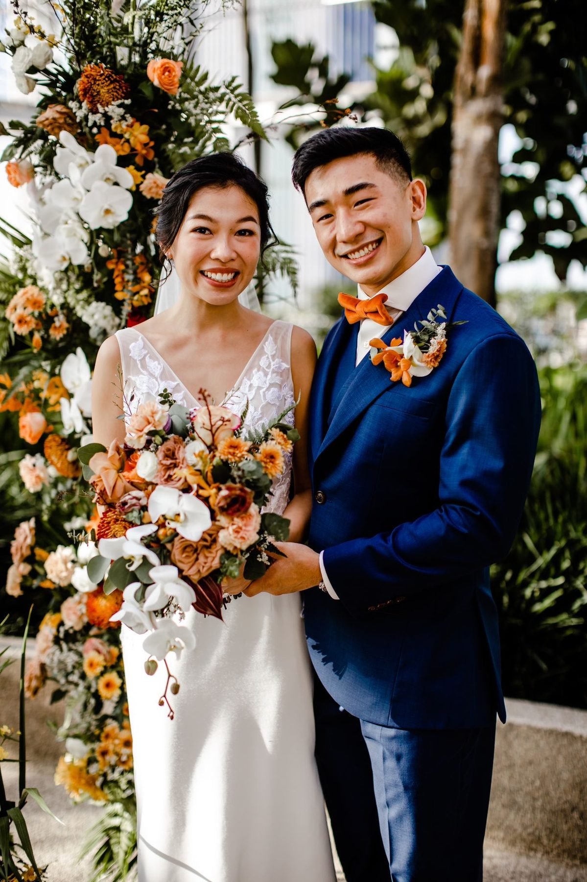 Bride in a white lace gown and groom in a navy suit with an orange bow tie smiling together in front of a tall orange and white floral pillar