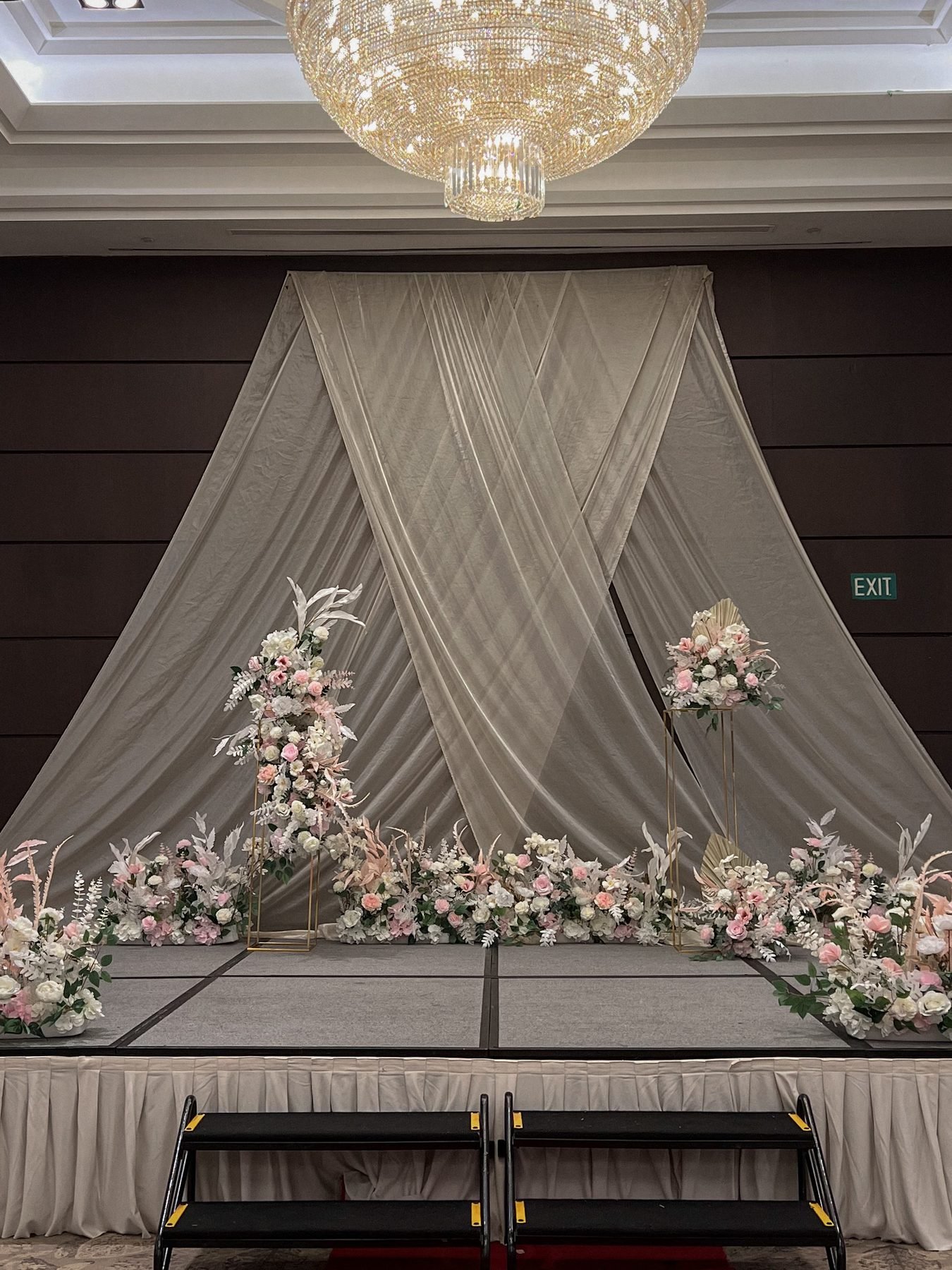A ballroom backdrop of draped champagne fabric with two gold pedestals of blush and white flowers