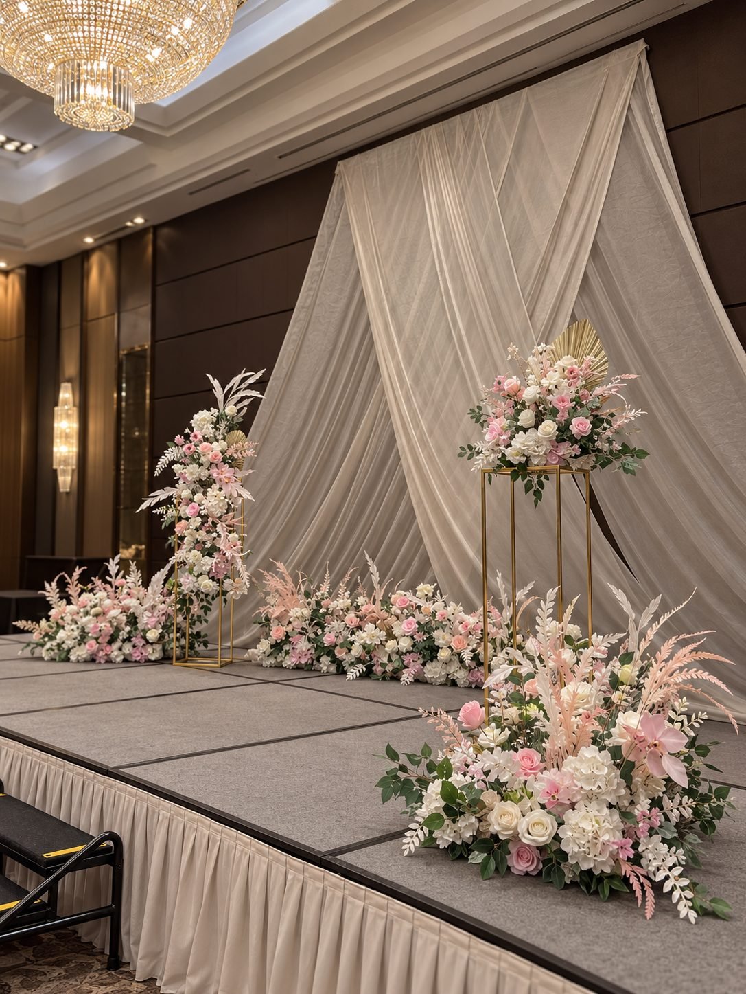 A ballroom stage with a criss-crossed sheer ivory drape backdrop, tall gold pedestals and low ground arrangements dressed in blush and white roses and pampas, beneath a crystal chandelier
