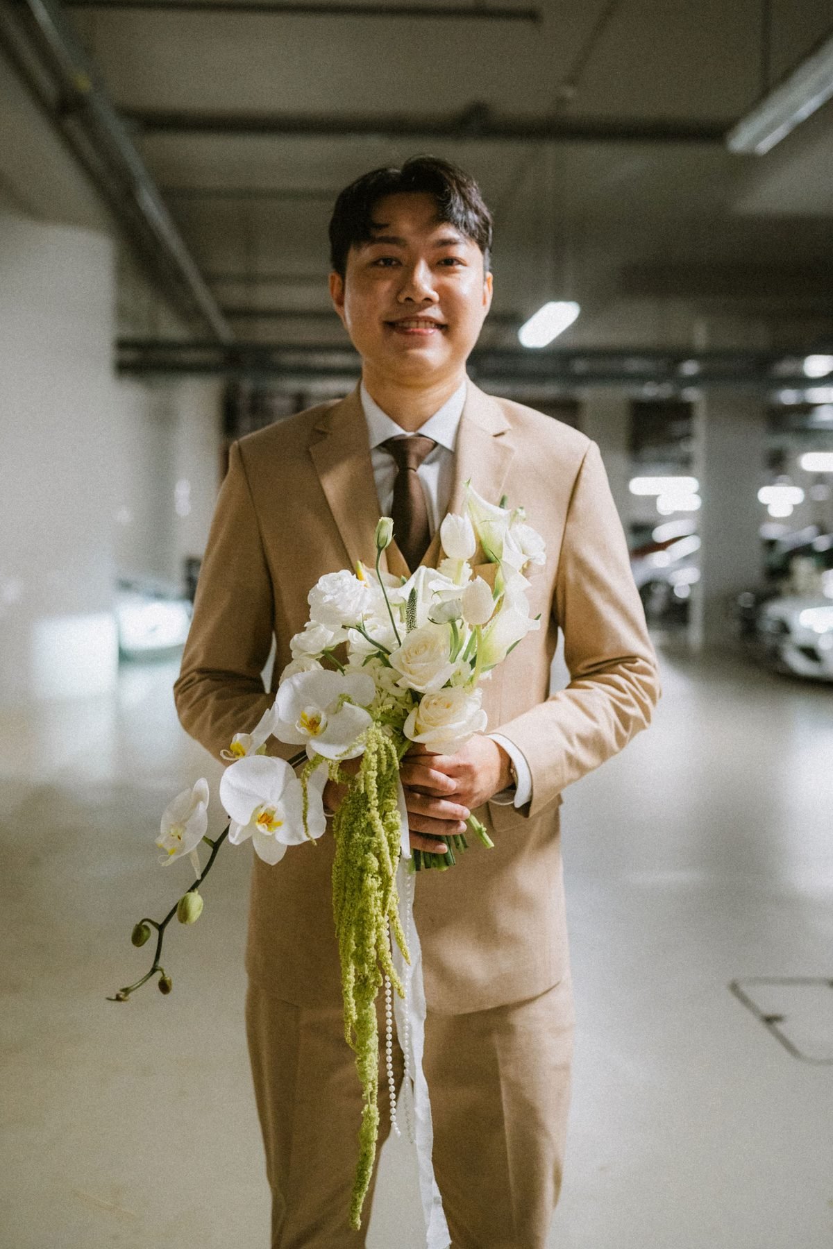 Groom in a khaki suit holding the long white cascading bouquet in an industrial corridor