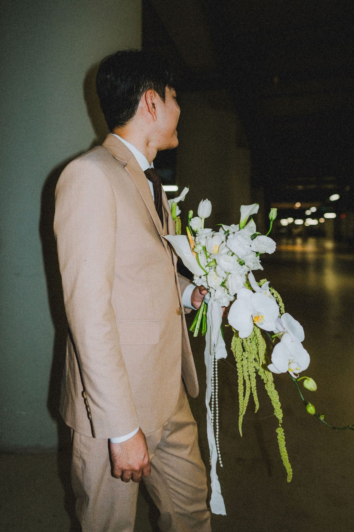 Groom in a khaki suit walking through a dark corridor while holding the long white cascading bouquet