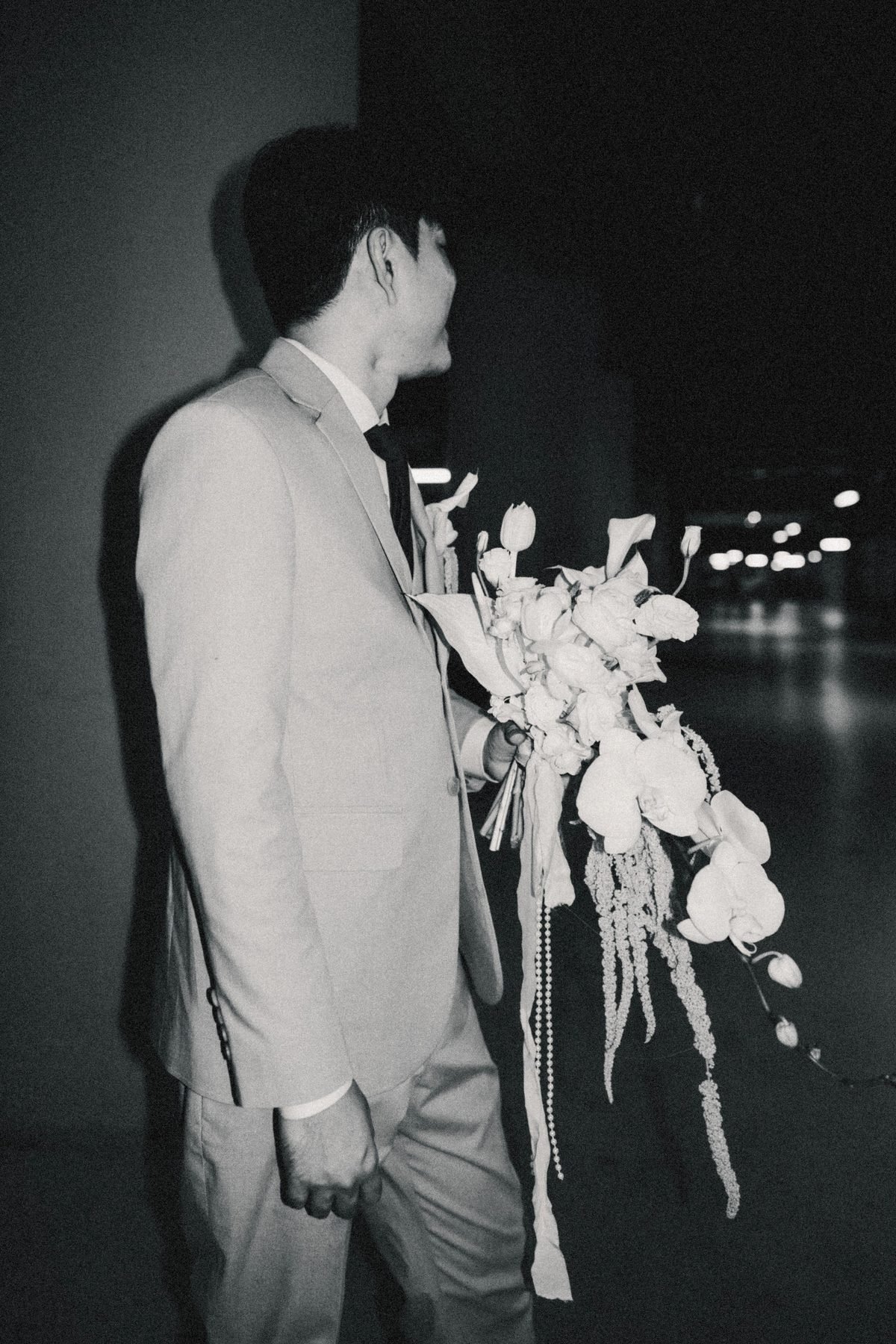 Black and white photograph of the groom in a khaki suit holding the cascading bouquet in a corridor