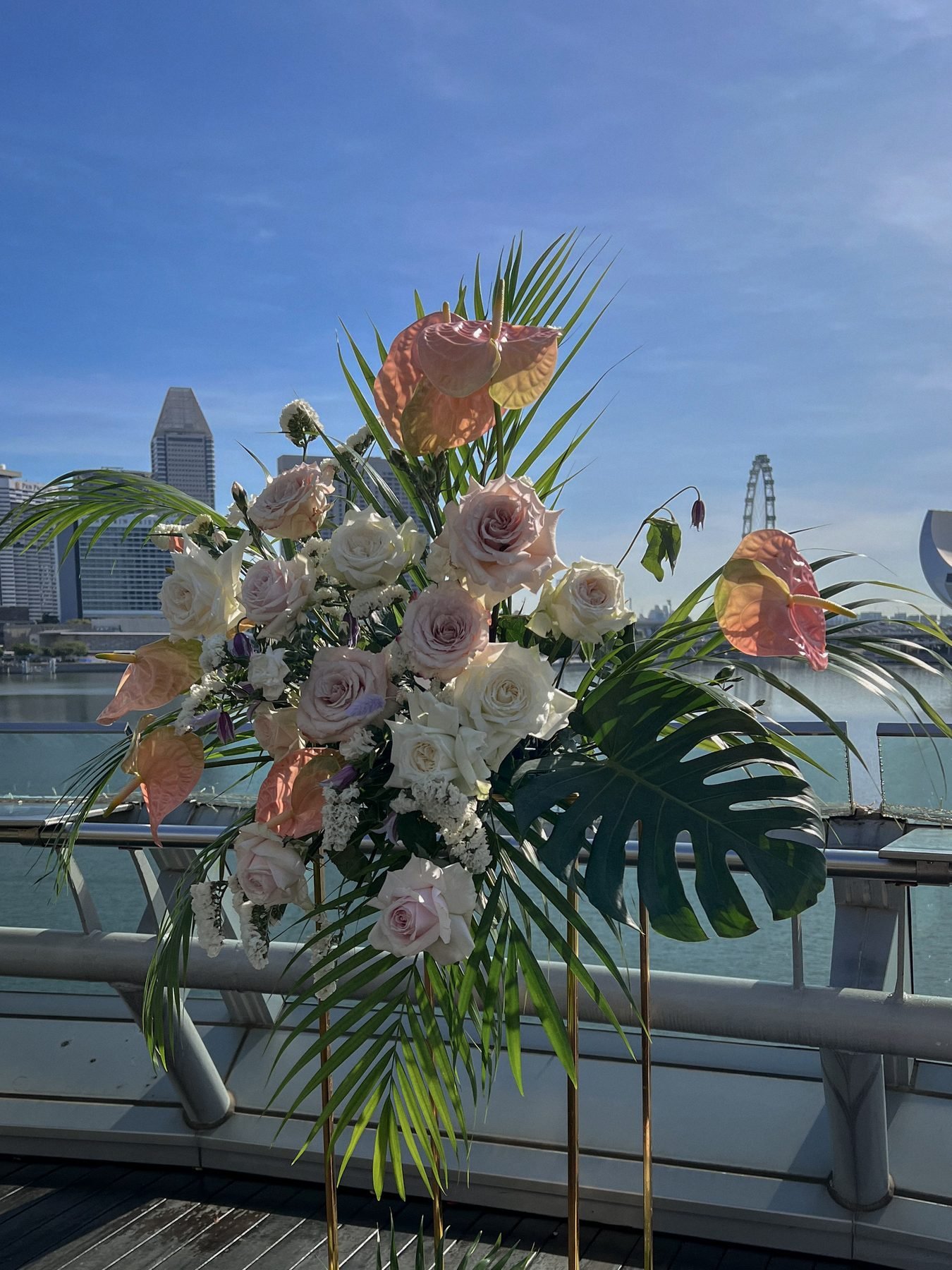 Tall standing arrangement of blush and peach anthuriums, roses and tropical leaves against a blue sky