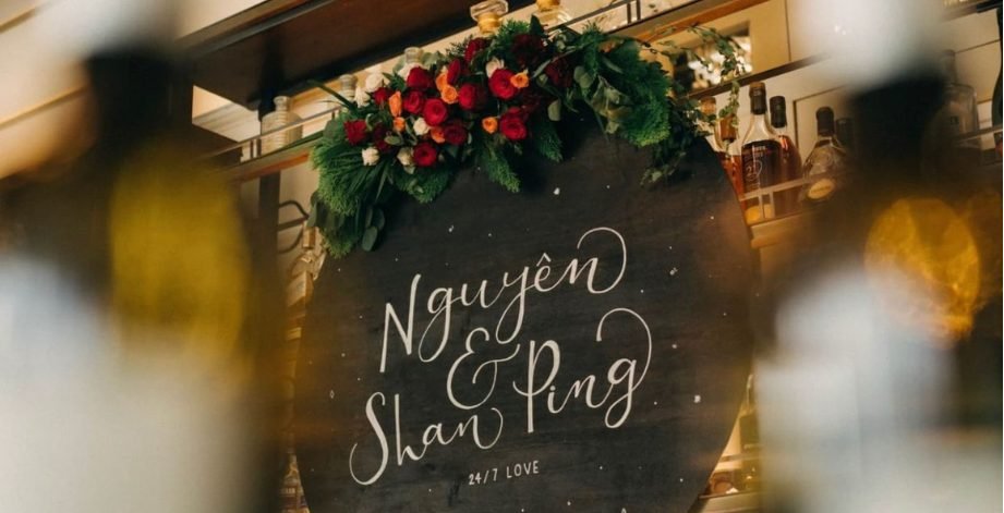 A dark round welcome sign hand-lettered with the couple names and date, topped with a swag of deep red, orange and white roses and greenery in front of a bar shelf of bottles