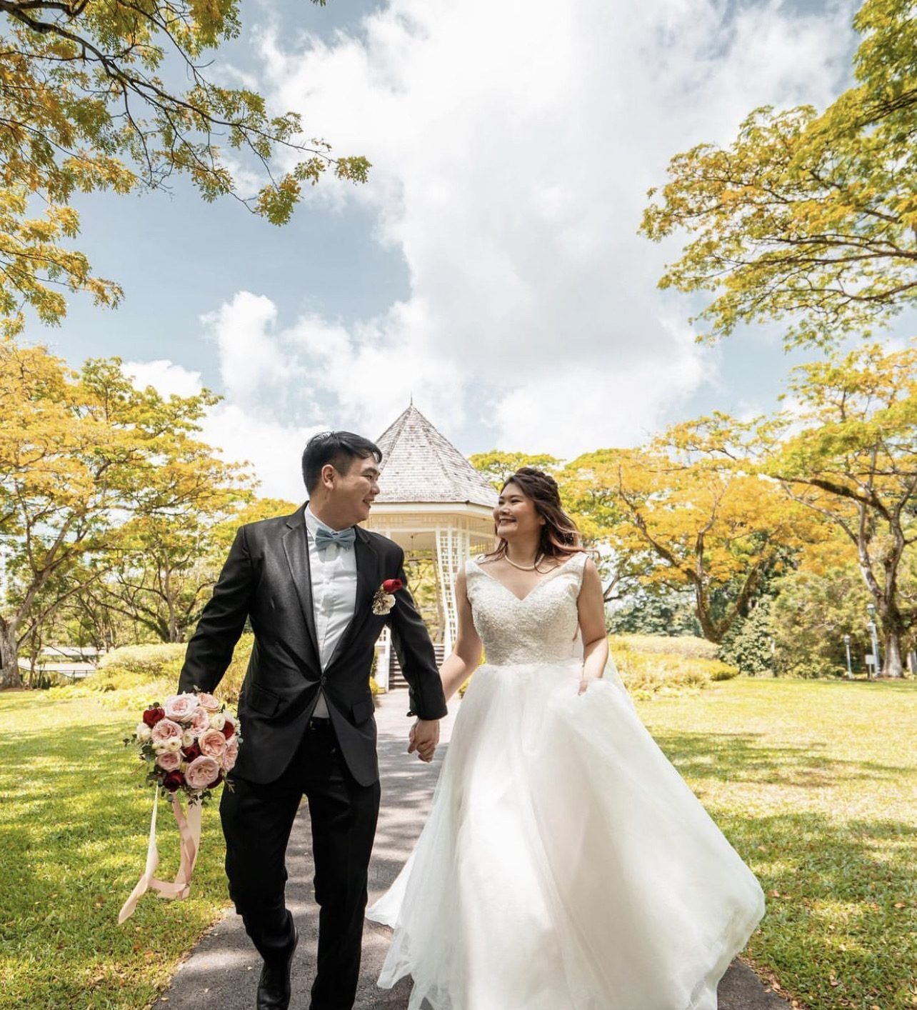 Groom in a black suit wearing a red boutonniere, holding a pink, red and ivory bridal bouquet with a trailing ribbon, walking hand in hand with the bride in a white dress through a park