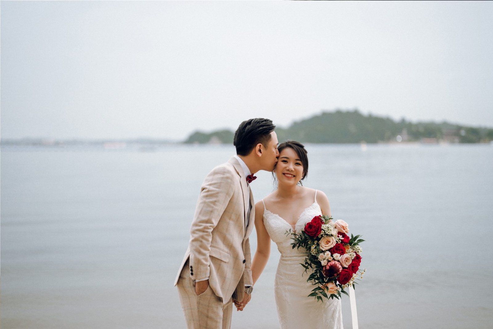 Groom in a beige suit kissing the bride's cheek by the water, her cascading red and pink bouquet in hand