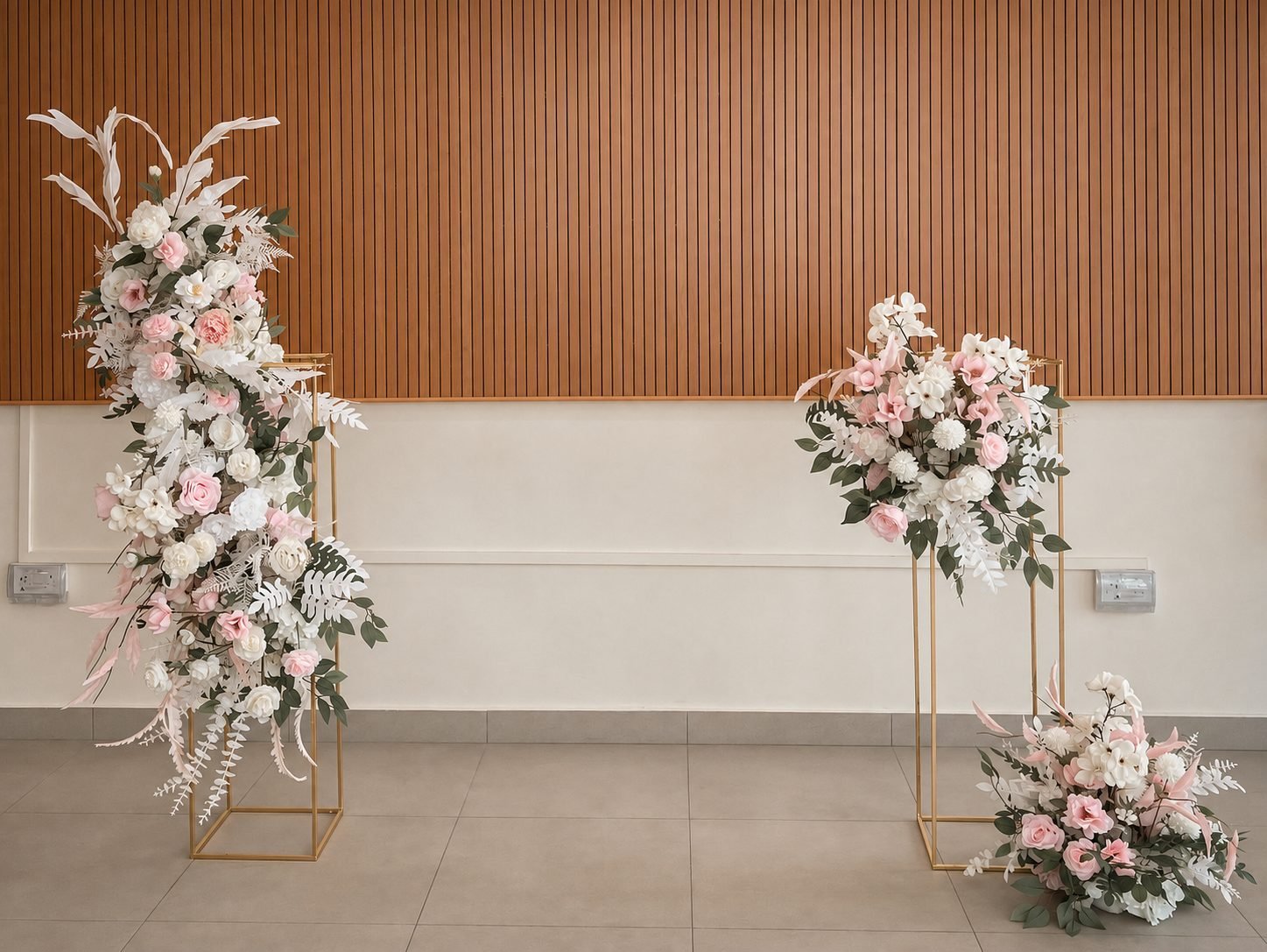 Blush and white floral arrangements on tall gold frames with a low ground arrangement against a wood-slatted wall