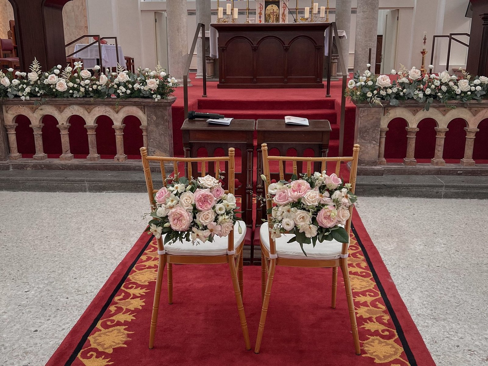 Two ceremony chairs with blush and white floral arrangements on the red-carpeted aisle, altar-rail flowers behind