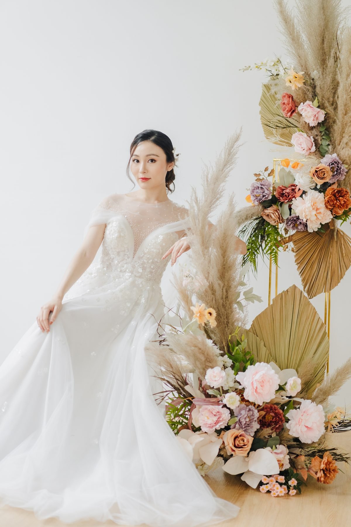 Bride in a white gown beside a tall pampas and dried-palm arrangement in blush and peach tones