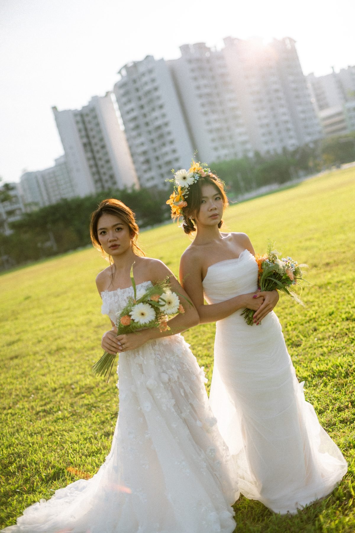 Two models in white gowns on a grass field holding small orange and white bouquets