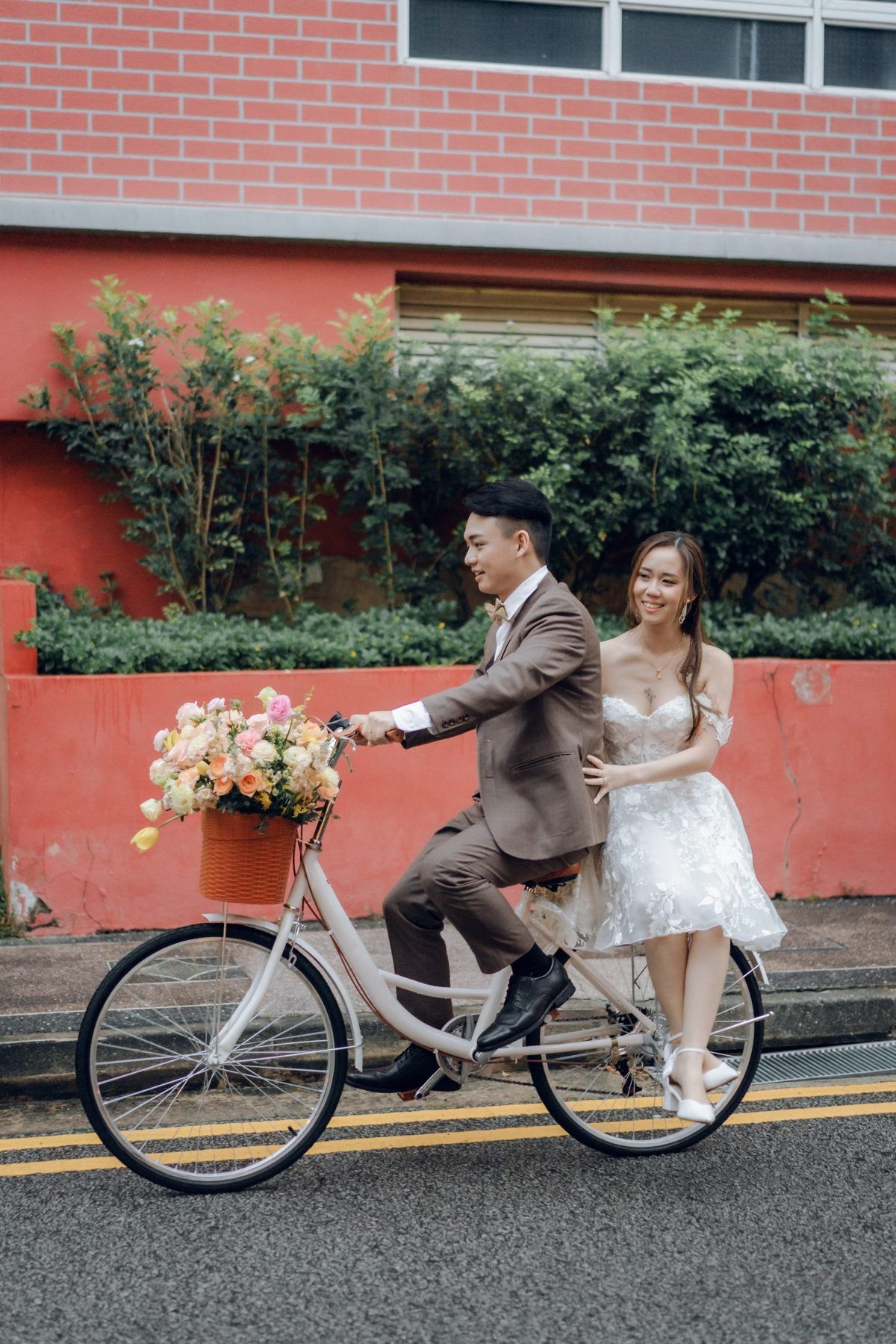 Couple riding a white bicycle with a flower-filled basket by a red brick wall