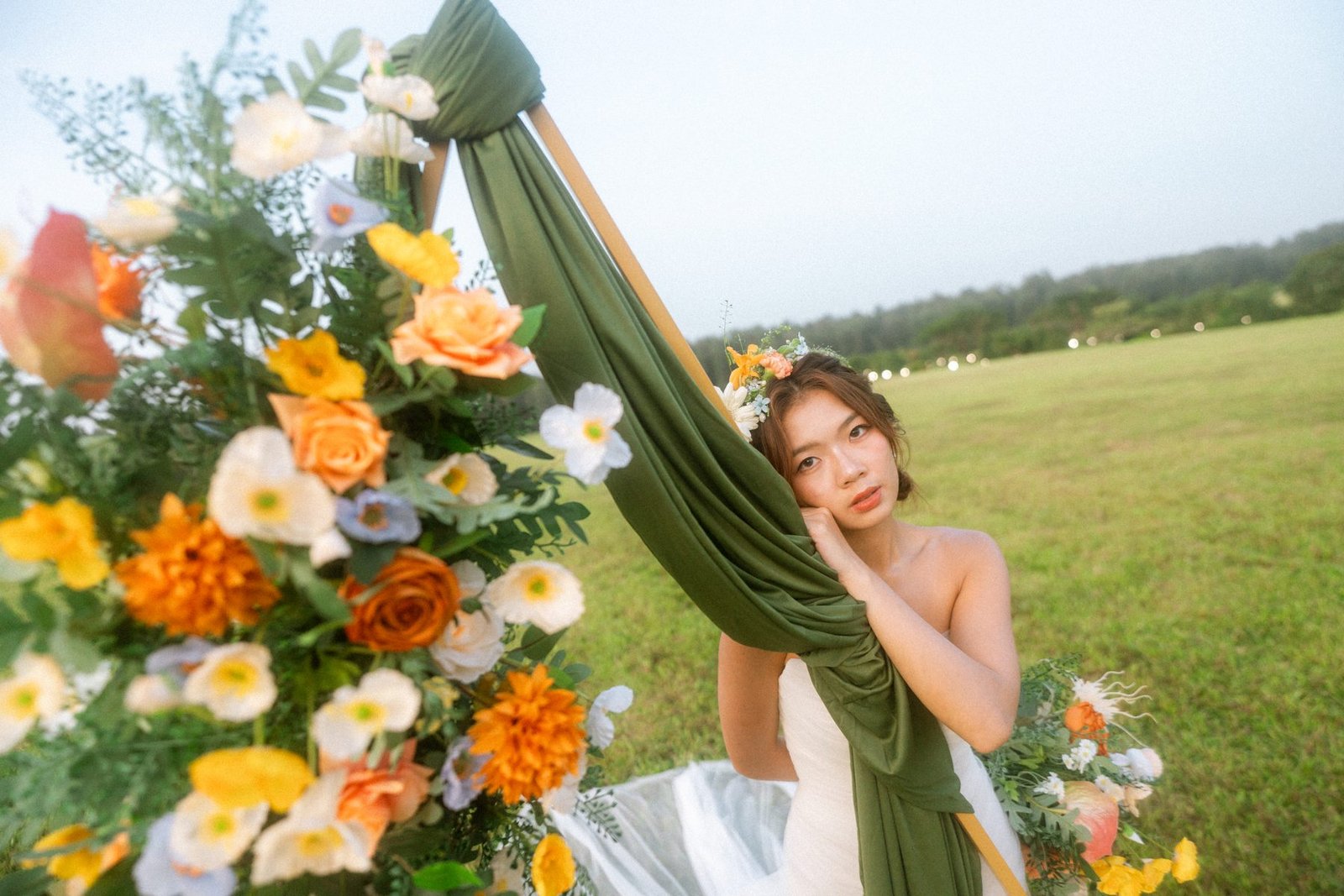 Bride leaning on an olive-draped arch beside a lush orange and white arrangement in a field